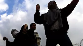 L'immagine mostra un gruppo di persone in una situazione di tensione, probabilmente in un contesto di protesta o conflitto. Alcuni individui sono coperti da sciarpe o fazzoletti, mentre sembrano lanciare dei proiettili, forse pietre. Sullo sfondo si possono vedere strutture architettoniche e un cielo nuvoloso, che contribuiscono all'atmosfera drammatica della scena. La presenza di filo spinato suggerisce una situazione di conflitto o di controllo militare.