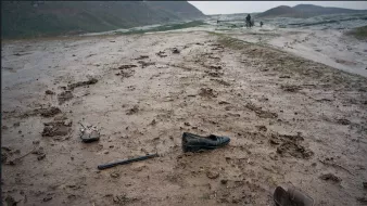 L'immagine mostra un paesaggio desolato, con un terreno fangoso e bagnato. Ci sono alcune scarpe abbandonate sulla strada, di cui una sembra essere un tacco alto e l'altra una scarpa più semplice. Sul suolo si notano segni di fango e detriti, creando un'atmosfera di degrado e abbandono. Sullo sfondo, si possono vedere delle figure lontane che camminano, suggerendo un contesto di difficoltà o di transito in un ambiente difficile. La scena comunica un senso di solitudine e trascuratezza.