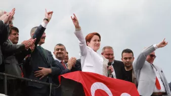 The image depicts a group of people at a political rally or event. In the foreground, a woman dressed in a white suit is speaking into a microphone, raising her arm with a gesture that signifies support or determination. Behind her, several men are visible, some clapping and others making similar gestures. The background features a large red banner, which may be associated with the event or political party. The overall atmosphere seems to convey enthusiasm and solidarity among the attendees.