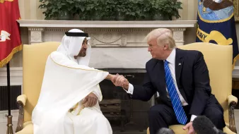 تظهر الصورة اجتماعاً بين رجلين يجلسان في غرفة تحتوي على تفاصيل فاخرة. الشخص في الجهة اليمنى يرتدي بدلة زرقاء مع ربطة عنق، بينما الشخص في الجهة اليسرى يرتدي الزي التقليدي الإماراتي. كلاهما يتصافح في لحظة ودية تعبر عن التعاون أو التفاهم. خلفهم توجد نباتات وزينة تتناسب مع أجواء الغرفة الرسمية.