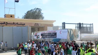 در تصویر، تعدادی از افراد در حال برگزاری یک تجمع یا اعتراض هستند. مردم در جلو ورودی یک ساختمان بزرگ ایستاده‌اند و برخی از آنها جلیقه‌های شرایطی به تن دارند. در پس‌زمینه، بنری بزرگ با متنی به زبان عربی و تصاویری از علم‌ها به چشم می‌خورد. همچنین در بالای این ساختمان، علم‌های کشورها در اهتزاز هستند. فضای تجمع به نظر شلوغ و پر انرژی می‌رسد.