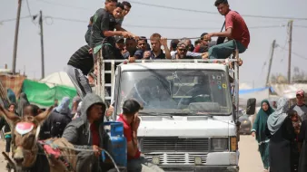 L'image montre un groupe de personnes assises sur le toit d'un camion, tandis que d'autres se trouvent à proximité, y compris des personnes à pied et une charrette tirée par un cheval. L'environnement semble être un endroit animé, avec des tentes et des structures visibles en arrière-plan. On peut voir des fils électriques et des montagnes de déchets, ce qui suggère une atmosphère informelle ou de camp. La scène dégage une impression de communauté et de vie quotidienne dans un contexte peut-être difficile.