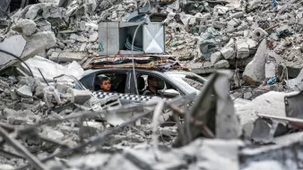 در تصویر، دو مرد درون یک خودرو در میان خرابه‌ها نشسته‌اند. اطراف آن‌ها پر از آوار و بقایای سازه‌های مخروبه است. برخی از أجزاء ساختمانی به وضوح دیده می‌شوند و وضعیتی به شدت آسیب‌دیده را نشان می‌دهند. در بالای خودرو، تجهیزات صوتی قرار گرفته است. فضای کلی تصویر حسی از ویرانی و بحران را منتقل می‌کند.