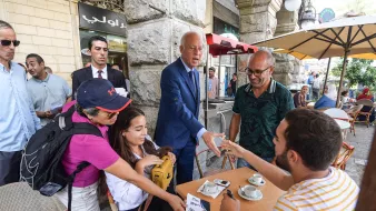 L'image montre une scène animée dans une rue. On peut voir un homme en costume, probablement une figure publique, en train de saluer des personnes autour de lui. Une femme avec un sac à dos et un chapeau semble lui tendre quelque chose. À la table, un groupe de personnes discute autour de tasses de café. L'atmosphère est conviviale et décontractée, avec des gens qui semblent engagés dans une conversation. Des parasols et des bâtiments historiques sont visibles en arrière-plan, ajoutant au cadre urbain.