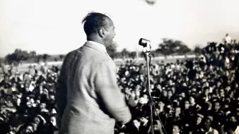 L'image montre un homme en train de parler sur une scène, devant un grand public. Il est vu de dos, et on peut voir une foule nombreuse devant lui, captivée par son discours. On distingue également un microphone sur le podium. L'atmosphère semble dynamique et engagée, probablement lors d'un événement politique ou d'un rassemblement. En arrière-plan, il y a des drapeaux, ce qui pourrait indiquer un contexte symbolique ou patriotique.
