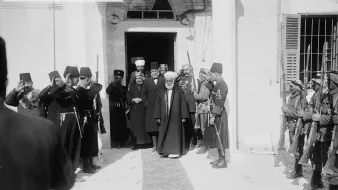 در تصویر، گروهی از مردان در یک مکان رسمی ایستاده‌اند. چند نفر از آنها در لباس‌های نظامی و دیگران در لباس‌های سنتی دیده می‌شوند. مردی با چهره‌ای جدی و کت و شلوار تیره در حال قدم گذاشتن به سمت دوربین است، در حالی که دیگران به او احترام می‌گذارند و سلام نظامی می‌دهند. محیط تصویر به نظر می‌رسد که یک ساختمان تاریخی یا دولتی باشد. فضای کلی نشان‌دهنده‌ی یک واقعه مهم و رسمی است.