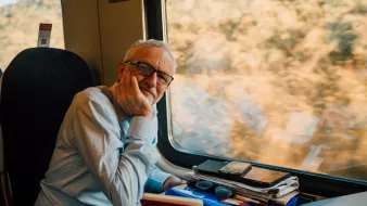 L'image montre un homme assis à côté d'une fenêtre de train. Il porte une chemise claire et a des lunettes. Son visage exprime une certaine réflexion ou contemplation alors qu'il repose sa tête sur sa main. Sur la table devant lui, on peut voir plusieurs objets comme des téléphones, des lunettes de soleil et des papiers, donnant l'impression qu'il a passé du temps à travailler ou à organiser ses affaires. À l'extérieur de la fenêtre, on aperçoit un paysage flou, suggérant que le train est en mouvement.
