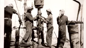 L'image représente un groupe de travailleurs en train de manipuler un équipement de forage, vraisemblablement sur un chantier pétrolier ou minier. Les ouvriers portent des vêtements de travail et des turbans, probablement pour se protéger de la poussière ou de la chaleur. Ils semblent concentrés sur leur tâche, travaillant ensemble sur une machine visible au centre de l'image. L'ambiance paraît industrialisée et intense, typique des travaux de forage au début du XXe siècle.