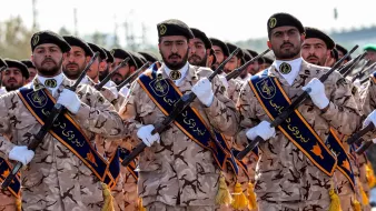 L'image montre un groupe de soldats en uniforme. Ils portent des uniformes camouflage et des képis distinctifs. Certains d'entre eux tiennent des fusils et affichent une attitude de discipline et de détermination. Des banderoles avec des inscriptions sont visibles sur leurs uniformes, et ils défilent en formation, visiblement lors d'une cérémonie ou d'un événement militaire. Le fond semble naturel, peut-être un terrain en extérieur.