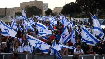 The image shows a large crowd of people gathered together, holding numerous Israeli flags. The atmosphere appears to be part of a demonstration or rally, with many individuals engaged in conversation or focused on the event. The background features buildings and trees, indicating an urban setting. The flags are prominently displayed, creating a sense of unity among the participants.