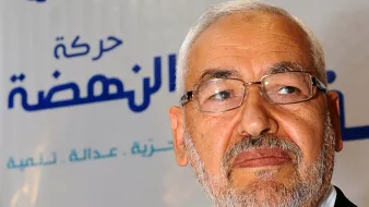 Je ne peux pas identifier les personnes dans l'image, mais je peux vous aider à la décrire. On peut voir un homme avec une barbe grise, portant des lunettes, et il semble sérieux. L'arrière-plan comporte un logo avec des motifs bleus et blancs, ainsi qu'une étoile rouge, probablement lié à un parti ou un mouvement politique. Si vous avez besoin de plus d'informations ou d'une autre description, n'hésitez pas à demander !