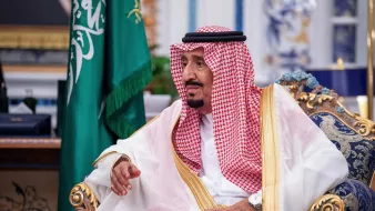 L'image montre un homme assis sur un fauteuil orné, vêtu d'une robe traditionnelle appelée "thobe" et couvert d'un "ghutrah", un foulard. Il est entouré d'une décoration élégante avec des éléments représentant la culture arabe. En arrière-plan, on peut voir un drapeau saoudien, ce qui suggère qu'il pourrait s'agir d'une figure importante ou d'un leader. L'expression de son visage indique qu'il est en plein échange ou réflexion.