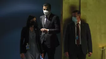 L'image montre trois personnes dans un hall. Deux d'entre elles portent des masques, et on peut voir qu'elles sont habillées de manière formelle. La personne à gauche semble avoir un badge autour du cou. Les trois individus se tiennent près d'un mur, avec une lumière dorée en arrière-plan. L'ambiance semble professionnelle et sérieuse.