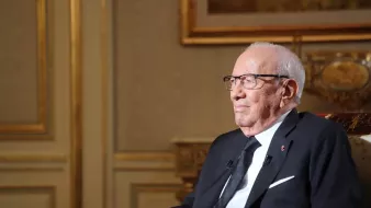 تظهر الصورة شخصًا مسنًا يجلس على كرسي في غرفة ذات ديكور فخم. يرتدي بدلة رسمية سوداء مع قميص أبيض وربطة عنق داكنة. يضع نظارات على عينيه ويبدو عليه التفكير والتركيز. خلفه توجد جدران مزخرفة وأمكنة تزينها الأعمال الفنية، مما يضيف لمسة من الأناقة إلى المشهد.