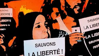 تظهر في الصورة مجموعة من الأشخاص المشاركين في احتجاج أو مظاهرة، حيث يحملون لافتات مكتوب عليها "لننقذ الحرية!" و"الحرية ستنقذ البقية...". يبدو من تعبيرات وجوههم أنهم متحمسون وملتزمون بالقضية التي يتظاهرون من أجلها. الخلفية تعكس أجواء حماسية ودعماً للقضية المطروحة.