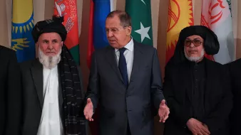 The image features a group of men standing in front of a backdrop adorned with various national flags. In the center is a man in a suit, possibly a diplomat or official, gesturing as he speaks. To his left and right are two men dressed in traditional attire, including a shalwar kameez and a shawl. The overall atmosphere suggests a formal meeting or diplomatic discussion, likely concerning regional issues or cooperation.