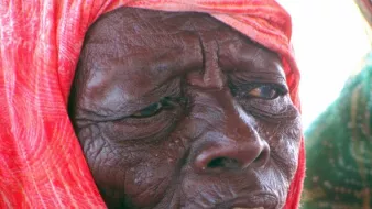L'image montre un portrait d'une personne âgée avec une expression réfléchie. Elle porte un foulard coloré, qui semble être enroulé autour de sa tête, et son visage présente des rides marquées, témoignant de son âge et de son vécu. La lumière met en valeur les détails de son visage.