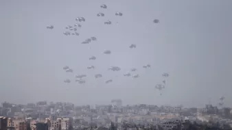 L'immagine mostra un cielo grigio-azzurro con numerosi paracadute che scendono lentamente verso un'area urbana sottostante. Sullo sfondo, è visibile una città con edifici di varie dimensioni e forme. L'atmosfera sembra tesa, suggerendo che si tratta di un'operazione militare o di emergenza. I paracadute, bianchi e leggeri, creano un contrasto visivo con l'ambiente circostante.