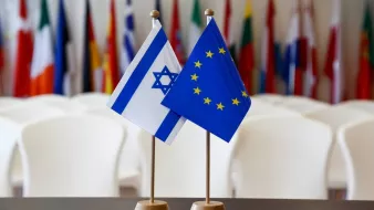 L'image montre deux drapeaux placés sur des supports. À gauche se trouve le drapeau d'Israël, qui présente une étoile de David et deux bandes bleues. À droite se trouve le drapeau de l'Union européenne, emblématique avec des étoiles dorées disposées en cercle sur un fond bleu. En arrière-plan, on aperçoit un rassemblement de drapeaux de différents pays, suggérant un contexte international ou diplomatique. Les chaises blanches sont disposées derrière, évoquant une salle de réunion ou une conférence.