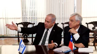 The image shows two men engaged in discussion at a table. One man is gesturing with his hand while speaking, suggesting he is making a point or explaining something. He is wearing a suit and a tie with stripes. The other man is sitting close by, listening attentively. He has gray hair and is also dressed in a suit. In front of them, there are flags of Israel and Chile on the table, indicating an official meeting or diplomatic discussion. The background features large windows with blinds, allowing natural light into the space.