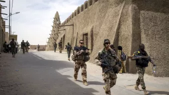 تظهر الصورة جنوداً يسيرون في شوارع منطقة تاريخية. المباني في الخلفية تبدو مصنوعة من الطين، وتمتاز بتصميم تقليدي. الجنود يحملون أسلحة ويبدو عليهم التركيز والحذر. يتواجد أيضاً شخصيات أخرى في الخلفية، مما يدل على وجود نشاطات متعددة في المنطقة. السماء تبدو صافية مع بعض السحب.