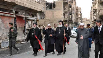 L'image montre un groupe de personnes marchant dans une rue endommagée. Il y a des bâtiments en ruines sur les côtés, indiquant une zone touchée par des conflits. Parmi le groupe, plusieurs hommes portent des vêtements traditionnels, notamment des robes noires et des croix, ce qui suggère qu'ils pourraient être des leaders religieux. Certains d'entre eux tiennent des bâtons. En arrière-plan, des militaires et des véhicules sont visibles, ajoutant à l'ambiance de la scène.