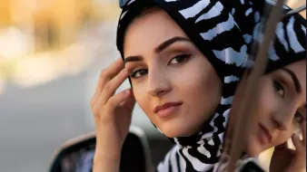 L'image montre une femme portant un hijab à motifs zebrés noirs et blancs. Elle se tient près d'une voiture, regardant vers la caméra avec une expression calme et confiante. Ses lunettes de soleil sont posées sur le haut de sa tête, ajoutant une touche de style. En arrière-plan, on peut apercevoir des contours flous d'un environnement urbain, suggérant un jour ensoleillé.