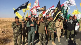 The image depicts a group of men participating in a march or rally, holding various flags. The men are dressed in military-style clothing and appear to be organized, with some wearing uniforms and others in casual attire. The flags they carry include a mix of colors and designs, suggesting they represent different factions or groups. The scene is set in an open area, likely with dry or arid terrain in the background, and the sky is clear. The atmosphere seems to indicate a demonstration or a show of solidarity among the participants.