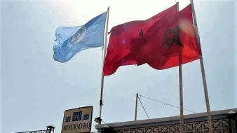 L'image montre deux drapeaux flottant sur des mâts. À gauche, il y a le drapeau de l'ONU, qui est de couleur bleu clair avec un emblème en blanc au centre. À droite, le drapeau du Maroc, qui est rouge avec une étoile verte à cinq branches. En dessous des drapeaux, on peut voir un panneau indiquant "MINURSO HQ", associé à des éléments graphiques de l'Union Européenne. L'ensemble de la scène est éclairé par une lumière vive, suggérant un temps ensoleillé.
