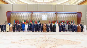 تظهر الصورة مجموعة من القادة ووزراء الخارجية المجتمعين في حدث رسمي. يقفون في صفوف مرتبة أمام خلفية مزينة بالعلم العربي، حيث تتواجد أعلام دول متعددة تشير إلى حدوث اجتماع أو قمة عربية. القادة يرتدون أزياء رسمية، بما في ذلك البذلات والعباءات، ويبدو عليهم الجدية والاهتمام بالمناسبة. الأجواء تعكس الطابع الرسمي والتاريخي لهذا اللقاء.