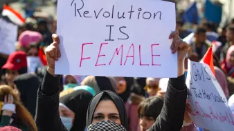 L'immagine mostra una manifestazione con molte persone riunite. In primo piano, una donna indossa un hijab e tiene in alto un cartello su cui è scritto "Revolution is Female". Sullo sfondo si vedono altre persone, alcune delle quali portano bandiere e cartelli, creando un'atmosfera di mobilitazione e protesta. La scena è caratterizzata da un forte senso di unità e determinazione.