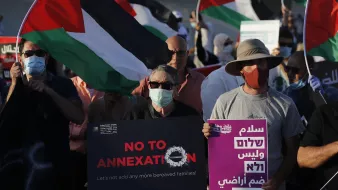 The image depicts a group of protesters holding flags and signs during a demonstration. The flags feature the colors of the Palestinian flag. Some participants are wearing masks, and a few signs read messages against annexation and calls for peace. The atmosphere appears to be one of solidarity and activism, as the individuals gather for a common cause.