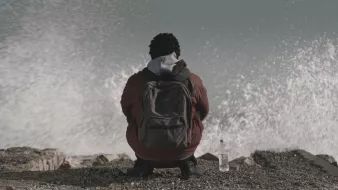 في الصورة، يظهر شخص يجلس على حافة شاطئ أو منطقة قريبة من البحر. يبدو أنه شاب ذو شعر مجعد، ويرتدي معطفًا سميكًا مع كيان داخلي ورأسه مائل قليلاً إلى الأمام. خلفه، تتلاطم أمواج البحر بقوة، مما يعطي إحساسًا بالحركة والطبيعة القوية. بجانبه، يوجد زجاجة ماء على الأرض. الأجواء تبدو هادئة بالرغم من قوة الأمواج، مما يضفي شعورًا بالتأمل أو الوحدة.