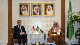 در تصویر، دو نفر در یک اتاق جلسه نشسته‌اند. در سمت چپ، یک مرد با کت و کراوات سیاه قرار دارد که به نظر می‌رسد فردی رسمی باشد. در سمت راست، مردی با لباس سنتی عربی نشسته است. پشت آن‌ها سه پرتره در قاب زرد رنگ قرار دارد که به نظر می‌رسد تصاویری از شخصیت‌های مهم هستند. همچنین، پرچم‌های ایالات متحده و عربستان سعودی روی میز قرار دارند و یک پرچم دیگر عربستان نیز در کنار آن‌ها دیده می‌شود. محیط اتاق با تزئینات مدرن و گیاهان تزئینی آراسته شده است.