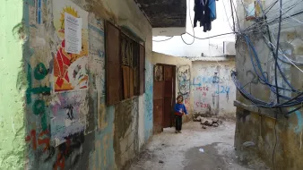 L'image montre une ruelle étroite et dégradée, bordée de murs en béton sur lesquels sont peints des graffitis et des affiches colorées. À gauche, une porte en bois est partiellement ouverte, tandis qu'à droite, on peut voir des fils électriques accrochés. Au fond de la ruelle, un jeune enfant se tient près de l'entrée, regardant vers l'objectif. L'atmosphère semble à la fois désolée et animée par les couleurs des murs et des affiches.