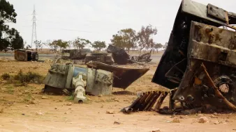 L'image montre un paysage désertique où sont dispersés des véhicules militaires rouillés et abandonnés. On peut voir des carcasses de tanks et d'autres machines, gisant sur le sol poussiéreux. Au loin, des arbres clairsemés et des pylônes électriques se distinguent, ajoutant à l'ambiance aride et désolée du lieu. Le ciel est nuageux, ce qui donne une atmosphère grise à la scène.