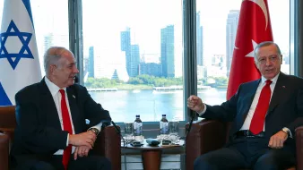 The image shows two men sitting in an office or meeting room with large windows that offer a view of a city skyline in the background. One man appears to be wearing a black suit and a red tie, while the other wears a dark suit and a white shirt with a red tie. There are symbols of Israel and Turkey nearby, indicating a diplomatic or official meeting between representatives of the two countries. The atmosphere seems cordial as they engage in conversation.
