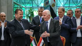 الصورة تظهر مجموعة من الشخصيات رجالية، تتواجد في مكان رسمي، حيث يتم عقد اتفاق أو توقيع. في المقدمة، يظهر شخصان shaking hands، يعبر ذلك عن اتفاق أو تعاون. الخلفية تحتوي على علمين: علم فلسطين وعلم مصر، مما يدل على موضوع النقاش أو الحدث. باقي الأشخاص في الصورة يبدون ممتنين أو يدعمون اللحظة، مما يعكس أهمية الحدث.