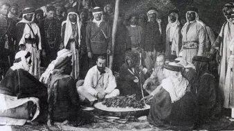 L'image montre un groupe de personnes, probablement issus de cultures arabes, rassemblées autour d'un grand plat de nourriture posé au sol. Certains sont assis tandis que d'autres se tiennent debout en arrière-plan. Les participants portent des vêtements traditionnels, tels que des robes et des turbans, ce qui suggère un contexte historique. L'ambiance semble conviviale, et l'événement pourrait être un repas ou une célébration. Des éléments du décor indiquent que la scène se déroule en extérieur, possiblement sous une tente.