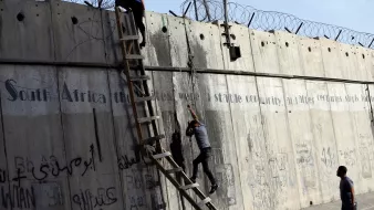 L'immagine mostra due persone che stanno scalando un muro di cemento, sul quale è presente della rete metallica. Dietro di loro, è visibile una scala che qualcuno sta utilizzando per salire. Il muro è coperto da graffiti e scritte, in varie lingue, che sembrano affrontare temi legati alla storia e alla condizione sociale. La scena evoca un senso di tensione e disperazione, con le persone che tentano di superare un ostacolo fisico, simbolo di divisione.