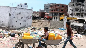 L'image montre deux enfants dans un environnement urbain en détérioration. L'un des enfants est assis dans une brouette, tandis que l'autre les pousse. Ils transportent des récipients jaunes, probablement pour l'eau, dans un paysage entouré de débris et de déchets. En arrière-plan, on peut voir des bâtiments en ruine et des camions. L'atmosphère semble difficile, reflétant une réalité de vie dans une zone touchée par des conflits ou des crises.