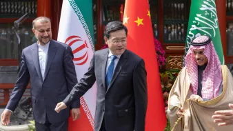 L'image montre trois hommes en costume, se tenant côte à côte devant des drapeaux. On devine que ces hommes représentent respectivement l'Iran, la Chine et l'Arabie Saoudite, compte tenu des drapeaux en arrière-plan. Ils semblent être en discussion lors d'un événement officiel. La scène est colorée, avec des éléments décoratifs comprenant des fleurs. Les hommes affichent des expressions sérieuses et engagées.