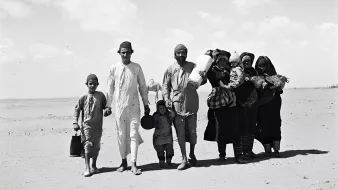 L'image montre un groupe de personnes marchant sur une surface sablonneuse, probablement dans un désert. Au premier plan, on peut voir plusieurs enfants et adultes. Les adultes portent des vêtements traditionnels. Certains membres du groupe transportent des objets sur leurs épaules ou dans les bras, tandis que les enfants semblent tenir la main de l'un des adultes. L'arrière-plan est vaste et dégagé, avec quelques nuages dans le ciel. L'atmosphère dégage une impression de déplacement ou de voyage.