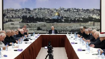 L'image montre une réunion se tenant autour d'une grande table ovale, avec plusieurs personnes assises de chaque côté. Au fond, il y a une grande image d'une ville, probablement Jérusalem, avec le dôme doré d'une mosquée bien visible. Les participants, qui semblent être des hommes âgés, portent des costumes sombres et affichent une expression sérieuse. Au centre de la table, une personne (probablement un chef ou un président) s'exprime pendant que les autres l'écoutent attentivement. Des bouteilles d'eau et des serviettes sont disposées sur la table. L'ambiance semble solennelle et attentive.