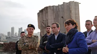 در این تصویر، گروهی از افراد در کنار یک ساختمان ویران، که ظاهراً بخشی از آن تخریب شده، ایستاده‌اند. یک نظامی با لباس نظامی و کلاه فرماندهی در سمت چپ قرار دارد. کنار او مردان و زنانی با لباس‌های رسمی و غیررسمی ایستاده‌اند و به نظر می‌رسد که در حال صحبت و تبادل نظر هستند. پس‌زمینه تصویر نشان‌دهنده شهری با ساختمان‌های بلند است. نور در تصویر کم است و فضای کلی حالت جدی و تاملی دارد.