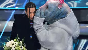 L'image montre une scène d'un spectacle où une personne au style vestimentaire unique, tenant un bouquet de fleurs, semble être en conversation avec une autre personne déguisée dans un costume original. Le costume ressemble à un animal aquatique, avec une tête d'apparence bleue et des détails colorés. L'ambiance semble dramatique ou émotive, avec un éclairage qui accentue les éléments de la scène.