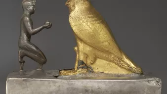 L'image représente une sculpture en métal, où l'on peut voir un personnage humain à genoux tenant un objet dans ses mains, face à un grand hibou doré. Le personnage est sculpté de manière détaillée, avec des traits qui évoquent un style ancien, probablement lié à l'Égypte ou à une culture similaire. Le hibou, de couleur dorée, symbolise peut-être la sagesse ou un aspect divin. La base de la sculpture est en métal argenté, offrant un contraste intéressant avec la brillance du hibou doré. Cette œuvre semble revêtir une signification symbolique ou religieuse.