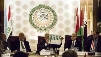 در این تصویر، گروهی از افراد در یک جلسه رسمی نشسته‌اند. یکی از افراد در حال نشان دادن یک نقشه یا نمودار به دیگران است. پشت آنها پرچم‌های کشورهای مختلف و نماد یا لوگوی اتحادیه عرب قابل مشاهده است. فضای جلسه حرفه‌ای و سازمان‌یافته به نظر می‌رسد. اعضای حاضر به دقت به موضوع مورد بحث گوش می‌دهند.