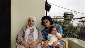 L'image montre trois femmes assises sur un balcon. À gauche, une femme âgée porte une robe grise et un foulard blanc. Au centre, une jeune femme, avec les cheveux noirs et bouclés, regarde vers l'objectif tout en tenant un enfant. L'enfant, assis sur les genoux de la jeune femme, semble jouer avec quelque chose. Le fond montre un environnement urbain avec des plantes et des éléments architecturaux. L'ambiance de l'image est chaleureuse et familiale.