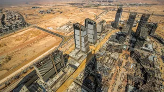The image showcases a panoramic view of a vast construction site in a desert landscape. In the foreground, several high-rise buildings are partially completed, with cranes and construction equipment evident. The surrounding area features empty land and some roadways, characteristic of a developing urban environment. The backdrop includes a clear sky and distant structures, suggesting a mixture of ongoing urbanization and natural desert terrain. The overall scene illustrates the rapid expansion of urban infrastructure in a typically arid region.